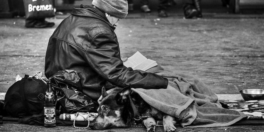 Schwarz-Weiß-Foto: Ein obdachloser Mann sitzt mit seinem Hund unter einer Decke in der Bremer Innenstadt auf dem Pflaster und liest ein Buch, im Hintergrund gehen Passanten vorbei.