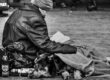Schwarz-Weiß-Foto: Ein obdachloser Mann sitzt mit seinem Hund unter einer Decke in der Bremer Innenstadt auf dem Pflaster und liest ein Buch, im Hintergrund gehen Passanten vorbei.
