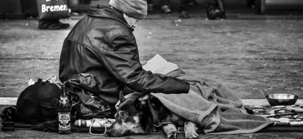 Schwarz-Weiß-Foto: Ein obdachloser Mann sitzt mit seinem Hund unter einer Decke in der Bremer Innenstadt auf dem Pflaster und liest ein Buch, im Hintergrund gehen Passanten vorbei.
